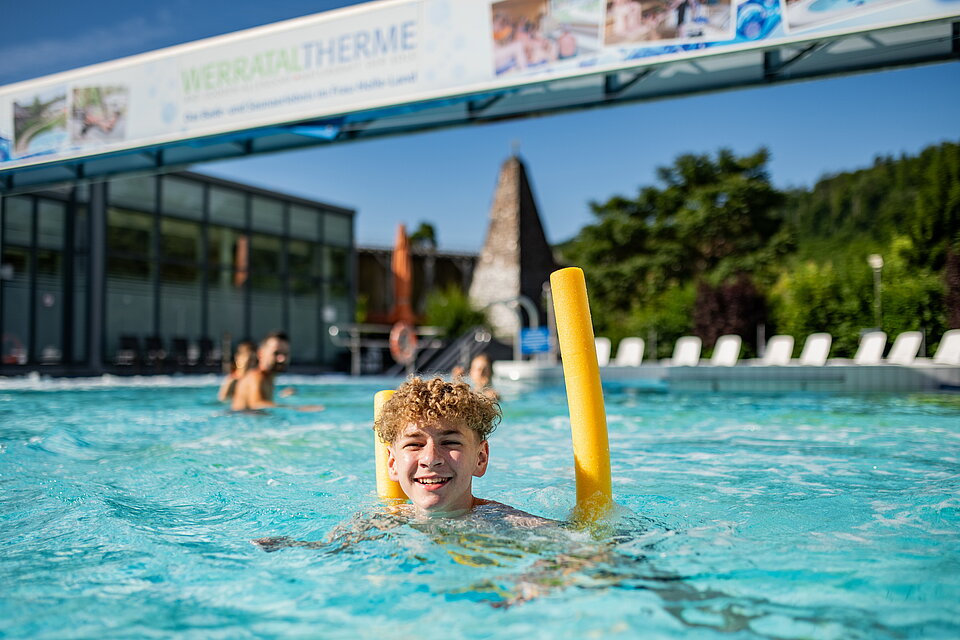Junge schwimmt mit einer gelben Poolnudel im Außenbecken.