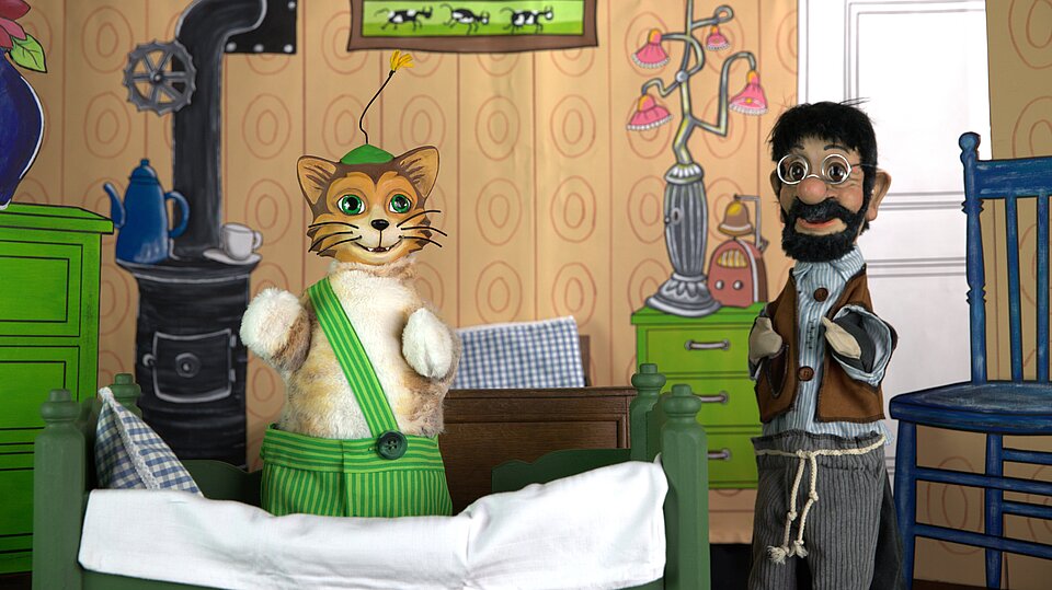 Ein rotgestreifter Kater mit grüner Latzhose sitzt auf einem Bett. Daneben steht ein Mann.