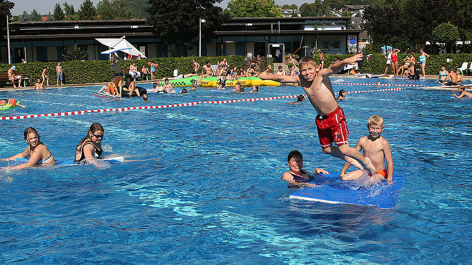 Spielende Kinder im Freibad. Ein Junge in roter Badehose springt von einer Luftmatratze ins Wasser