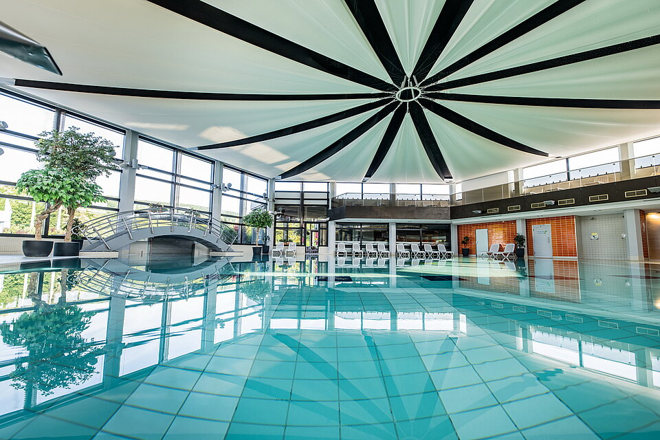 Großes Schwimmbecken mit klarem Wasser. An der Seite stehen weiße Liegestühle.